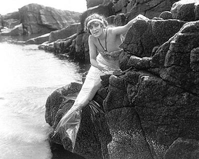 Annette Kellerman, in mermaid costume in role of &quot;Merrilla, the Queen of the Sea&quot;, looks around some rocks on a sea-shore. From 1918 film &quot;Queen of the Sea.&quot;