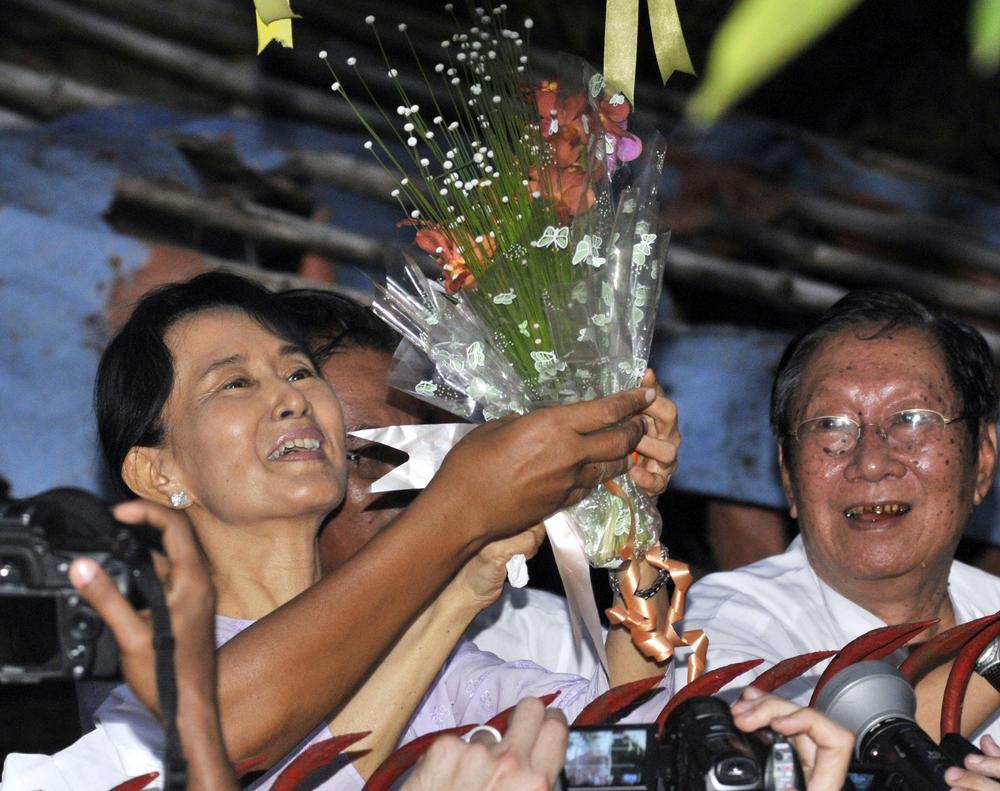 Myanmar&#39;s pro-democracy leader Aung San Suu Kyi, left, smiles after she received flowers from her supporters as she stands at the gate of her home Saturday. (AP)
