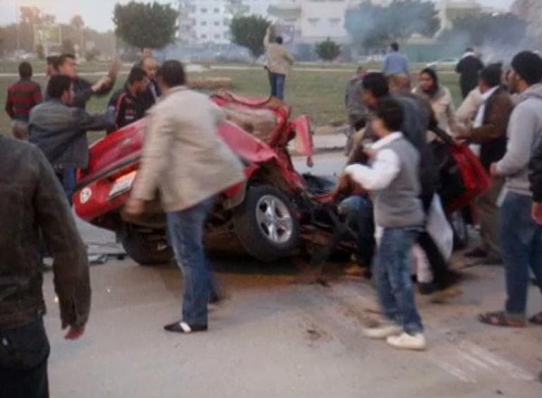 This image taken from amateur video obtained Thursday Feb. 24, 2011 and released by Libyan opposition groups, purportedly shows a crushed red car surrounded by a gesticulating and distressed crowd, as tanks roll in the distance releasing exhaust smoke between Feb 18-19, 2011 in Benghazi, Libya. (AP Photo via APTN) 