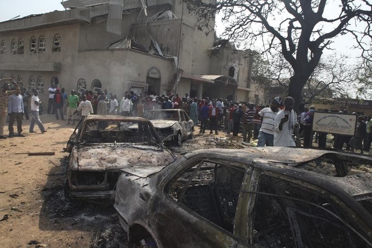 Onlookers gather around a destroyed car at the site of a bomb blast at St. Theresa Catholic Church in Madalla, Nigeria. (AP)
