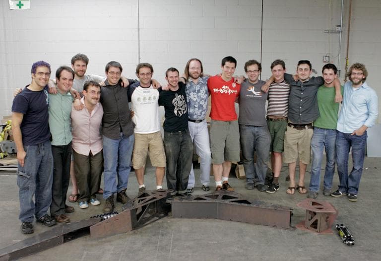 The Project Hexapod class, consisting of 3 instructors, 1 TA, and 15 students standing by a scale model of Stompy's leg (Nate Goldman/WBUR)