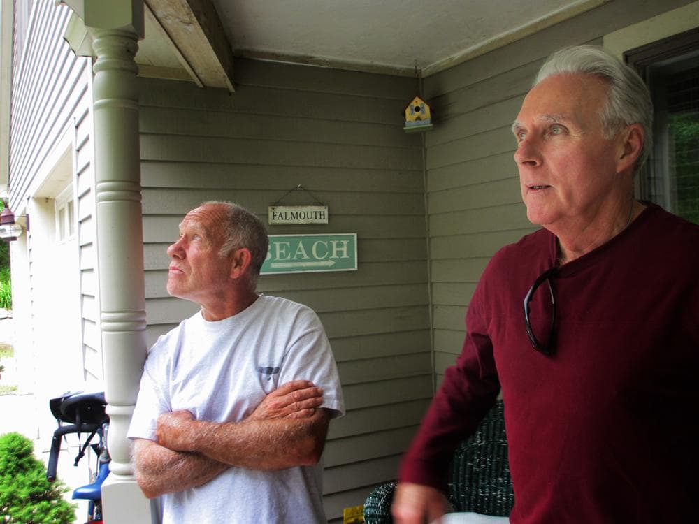 Neil Andersen and John Ford stand on the back porch of Andersen’s house on Blacksmith Shop Road in Falmouth. Both say the town&#039;s wind turbines have caused them health issues. (Kathleen McNerney/WBUR)