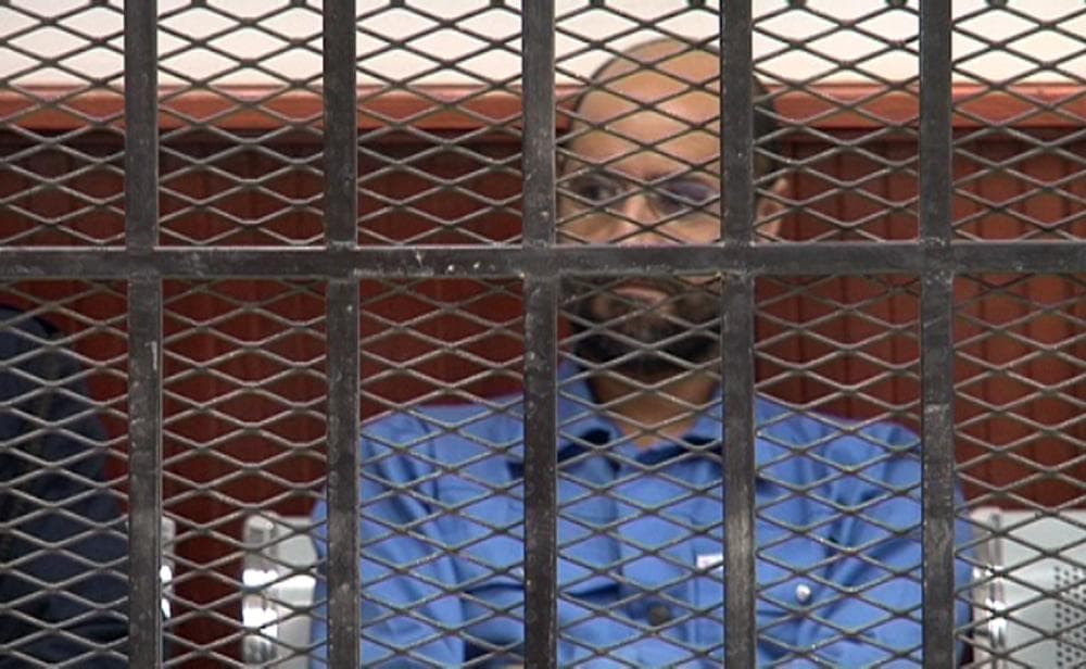 This image made from video distributed by the Zintan Media Center shows Seif al-Islam Gadhafi, center, inside a defendant's cage in a courtroom in Zintan, Libya, Thursday, May 2, 2013. (Zintan Media Center via AP)