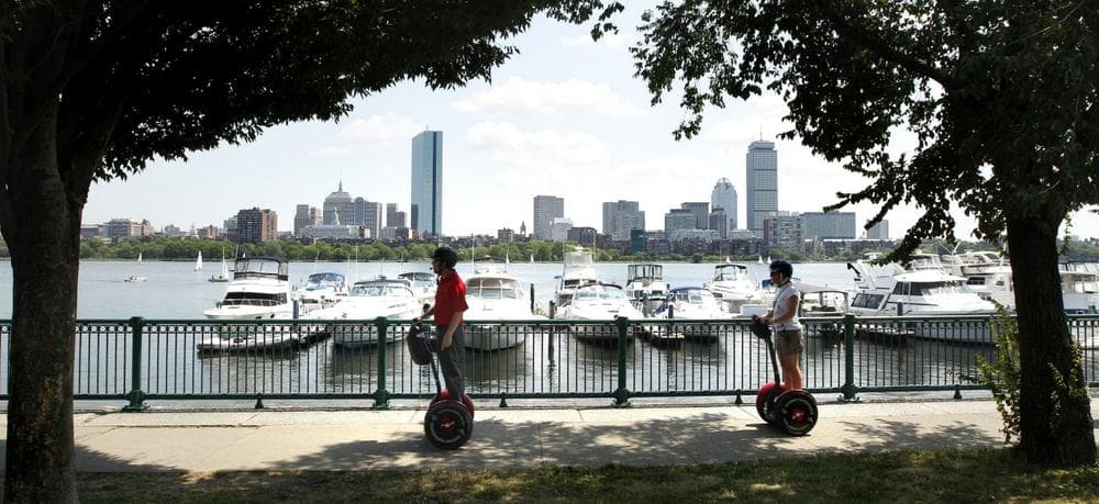 With the Boston skyline in the background, riders roll down a sidewalk on Segways along the Charles River in Cambridge, Mass. (AP)