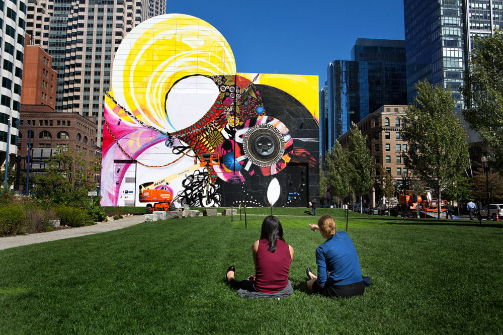 Two women sitting on the grass during a lunch break point out elements of the new mural at Dewey Sq.  (Jesse Costa/WBUR)