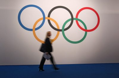 A woman walks in front of a board showing Olympics rings at the 127th International Olympic Committee session in Monaco, Monday, Dec. 8, 2014. (AP)