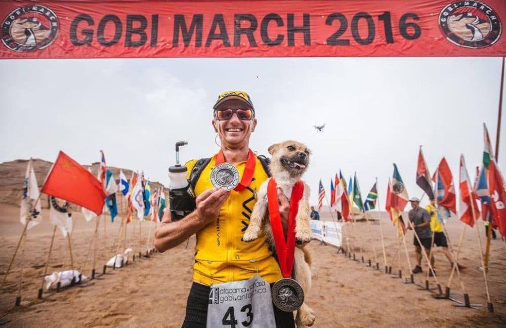Scottish ultramarathoner Dion Leonard stands at the finish line of the Gobi March in China, holding Gobi, a stray dog who ran alongside him for most of the 155-mile race. (Courtesy of Bring Gobi Home via Facebook)