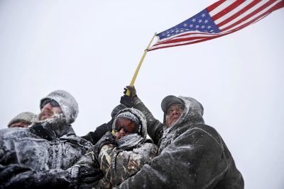 Military veterans huddle together to hold a United States flag against strong winds during a march to a closed bridge outside the Oceti Sakowin camp where people have gathered to protest the Dakota Access oil pipeline in Cannon Ball, N.D., Monday, Dec. 5, 2016. (David Goldman/AP)