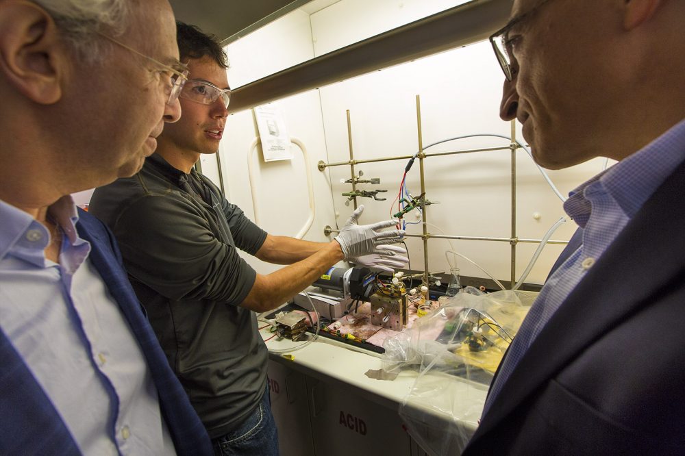 Harvard graduate student Andrew Wong, center, demonstrates his lab prototype of an organic mega flow battery to professor Roy Gordon, left, and professor Michael Aziz. (Jesse Costa/WBUR)