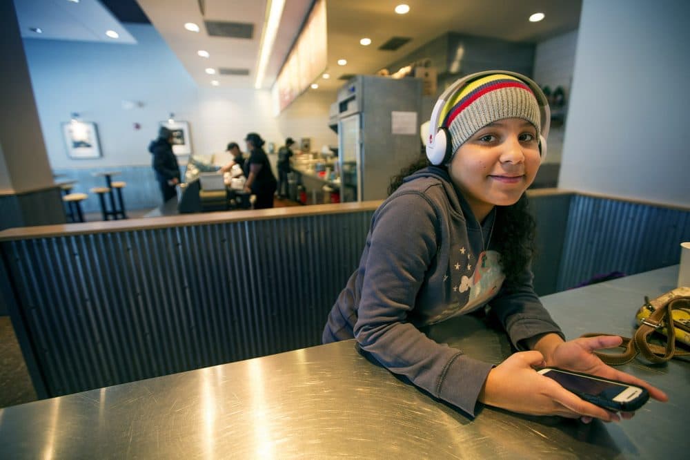 During her day off from school, Xiomary Gonzalez, 10, plays White Tiles 4 on her phone to pass the time away while her mother, Xiomara, works at the register at Chipotle. (Jesse Costa/WBUR)