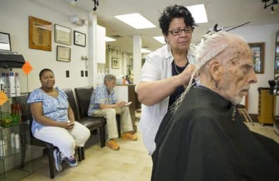 Caretaker Yamiley Jean Louis looks on as Leroy Neuberg, 102, gets his regular haircut from Maria Chavez at Salon Maria in Brookline. (Robin Lubbock/WBUR)