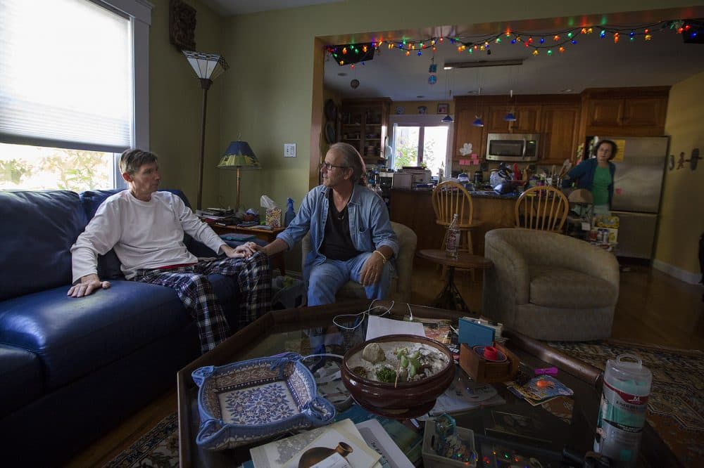 Ron Hoffman, center, and Don Viewegtalk, who has ALS, talk in the living room while Don's wife Dana prepares his lunch. He needs a feeding tube to eat. (Jesse Costa/WBUR)