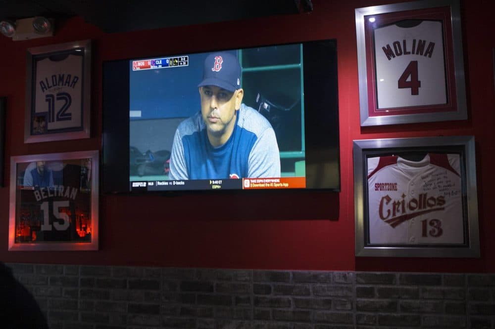 Red Sox manager Alex Cora is seen on a TV in his hometown of Caguas, Puerto Rico, during a game against the Indians. On the wall around the TV are other baseball players' jerseys. (Jesse Costa/WBUR)