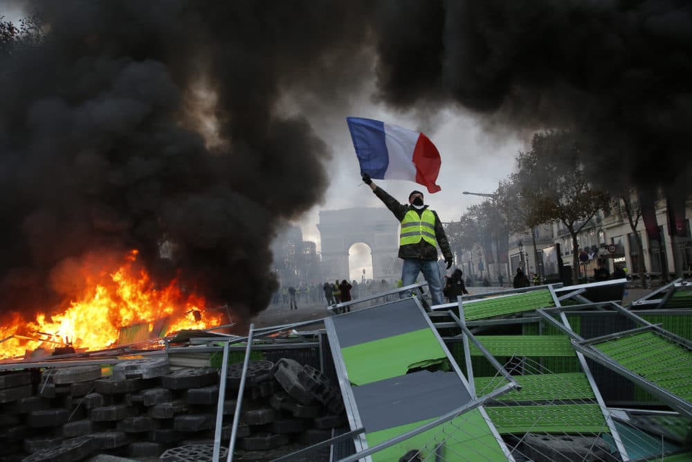A demonstrator waves the French flag onto a burning barricade on the Champs-Elysees avenue during a demonstration against the rising of the fuel taxes, Saturday, Nov. 24, 2018 in Paris. French police fired tear gas and water cannons to disperse demonstrators in Paris Saturday, as thousands gathered in the capital and staged road blockades across the nation to vent anger against rising fuel taxes and Emmanuel Macron's presidency. (Michel Euler/AP)