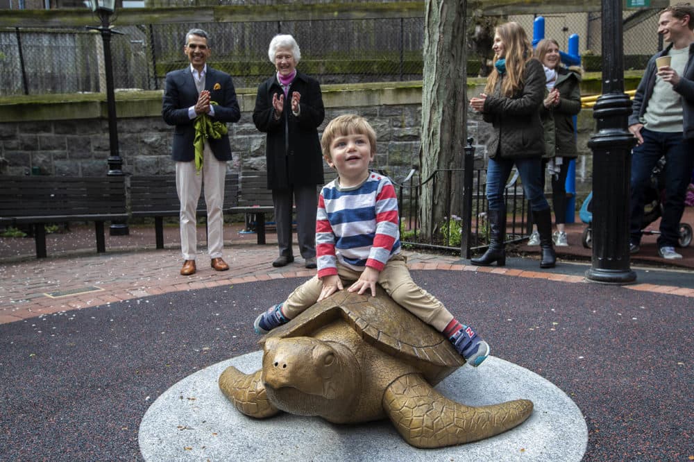 Sculptor Nancy Schön, right, and Miguel Rosales, left, applaud as 2-year-old Winston Leffler is the first child to climb aboard “Myrtle The Turtle” at the Myrtle Street Playground in Beacon Hill. (Jesse Costa/WBUR)