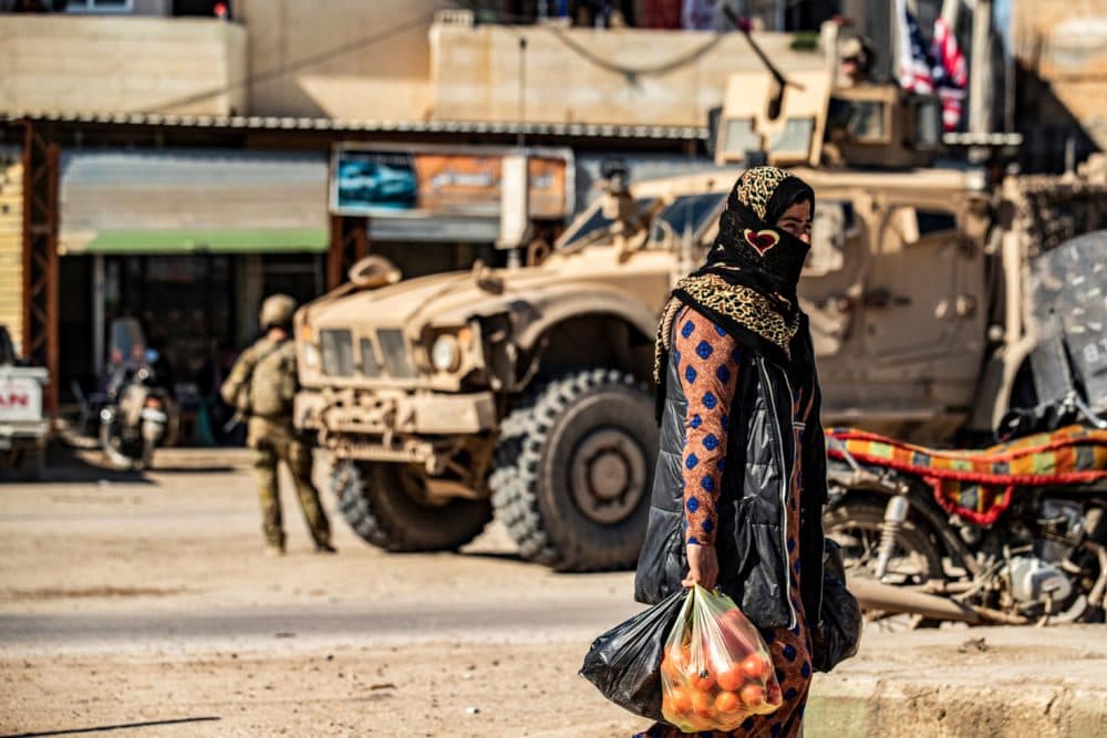 A woman carrying produce walks past a US military armoured vehicle in the town of Tal Tamr along the M4 highway in the northeastern Syrian Hasakeh province, near the border with Turkey, on March 3, 2020. (DELIL SOULEIMAN/AFP via Getty Images)