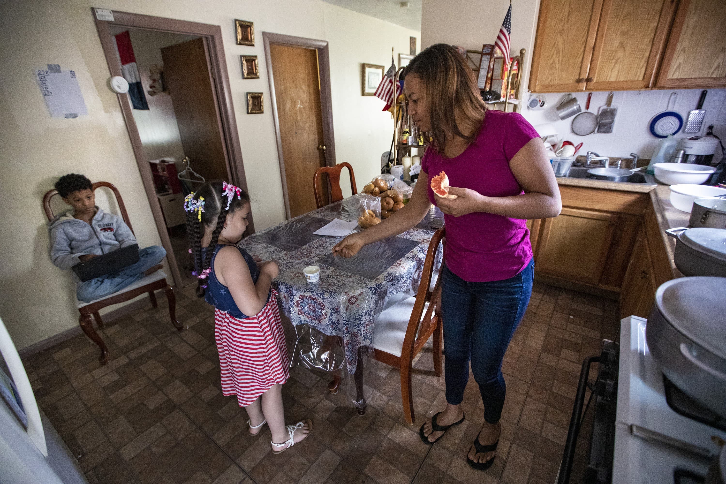 Margareth gives a spoon to Luz to eat her late morning yogurt snack, while Jesus sits on a chair using a laptop. (Jesse Costa/WBUR)
