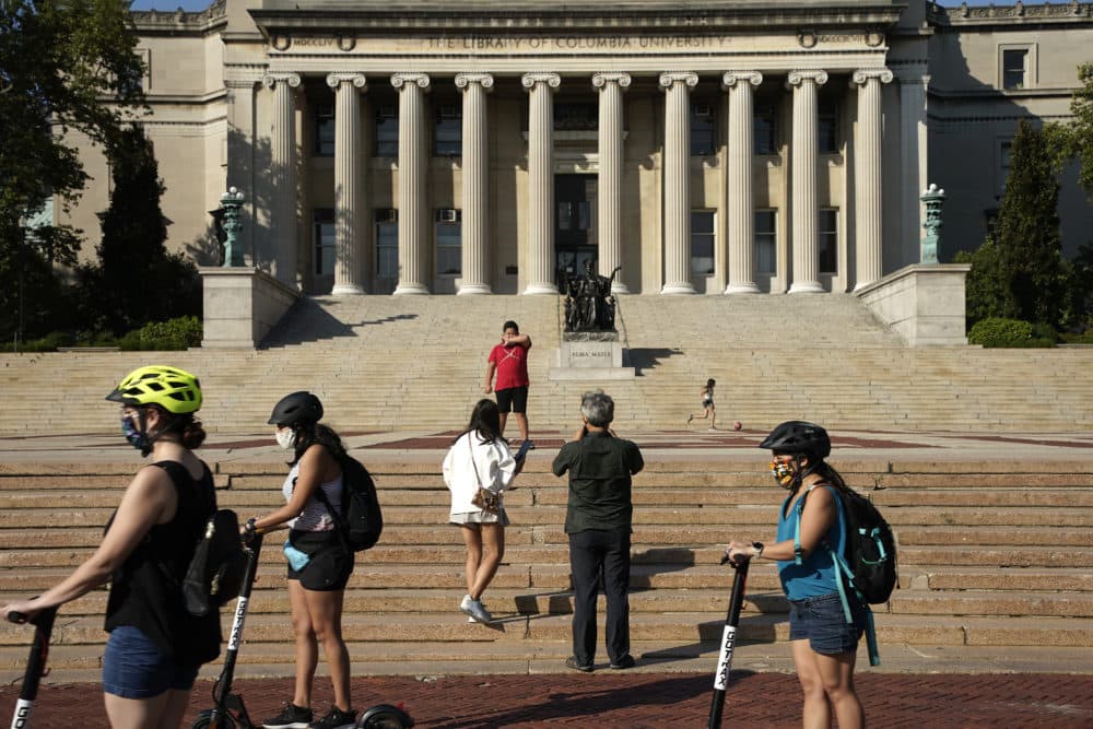 People wearing masks ride electric scooters as visitors take photos in front of Columbia University's Low Memorial Library on August 23, 2020 in New York City.  (John Lamparski/Getty Images)