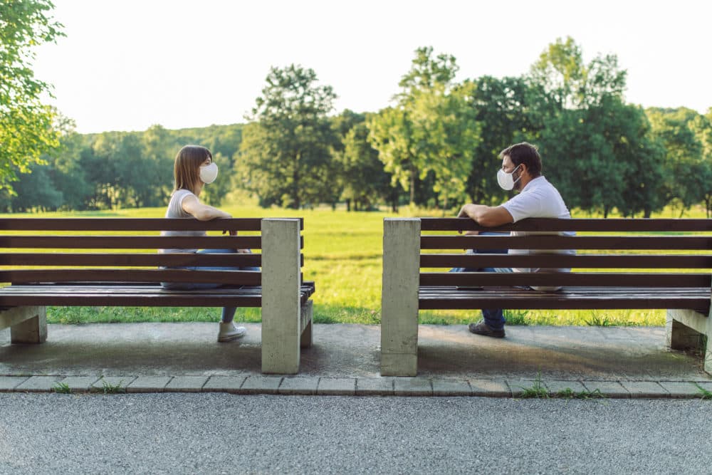 Two friends maintain social distance by sitting on separate benches and wearing protective face masks. (Getty)
