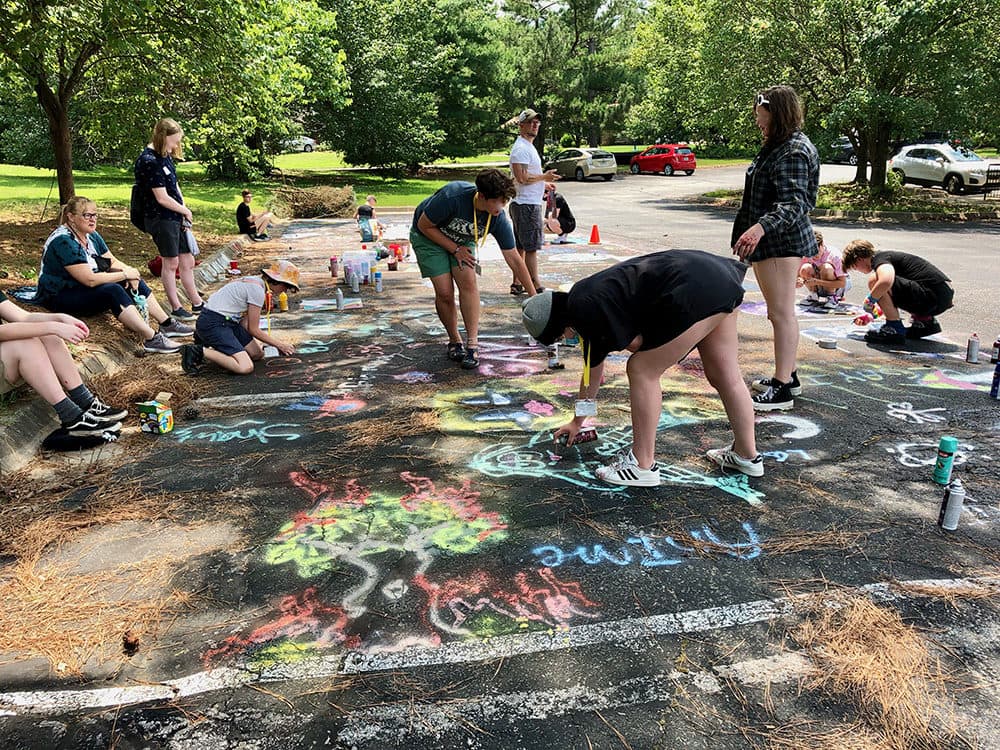 Queer camp youth ages 12 to 18 spray paint and glitter artworks on the parking lot of Good Shepherd Lutheran Church as parents and a camp counselor observe on July 6, 2021, in Fayetteville, Arkansas. (Jacqueline Froelich/KUAF)