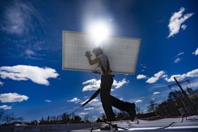 A technician walks with a solar panel across the rooftop during a solar panel installation at Boston Building Resources in Jamaica Plain. (Jesse Costa/WBUR)