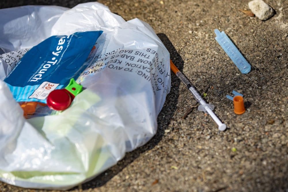 A syringe filled with what is believed to fentanyl lies on the ground with a bag of other various drug user paraphernalia behind the Universal Missionary Church in Brockton. (Jesse Costa/WBUR)
