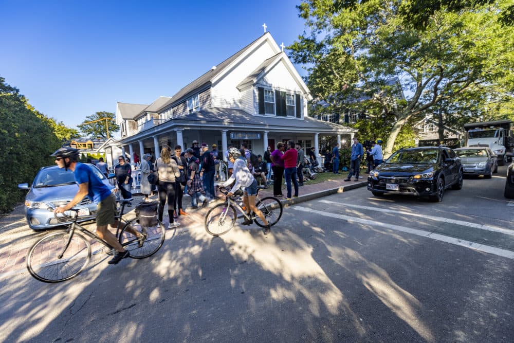 Cyclists and drivers of automobiles pass by the scene at St. Andrew’s Parish House in Edgartown where dozens of migrants from Venezuela were transported from the South to Martha Vineyard. (Jesse Costa/WBUR)