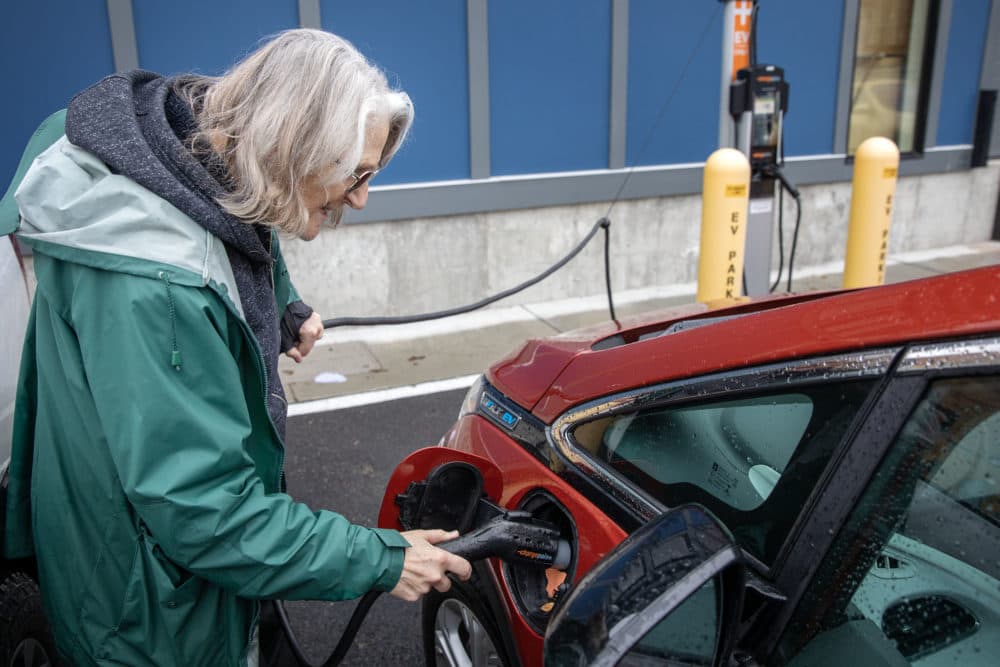 Loie Hayes plugs in her car at a charging station in Boston. She wears glasses and a green rain coat, she has white hair. Her car is a red electric car. (Credit: Robin Lubbock/WBUR)