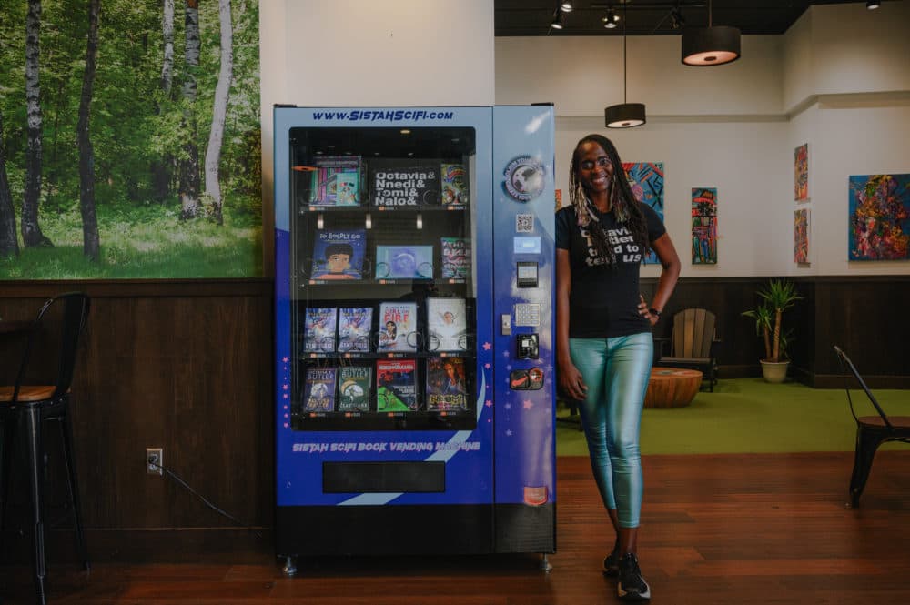 Sistah Scifi founder Isis Asare stands for a portrait next to her new book vending machine inside Oaklandia Cafe x Bakery in Downtown Oakland on February 22, 2023. Asare hopes to create a national network of Sistah Scifi vending machines to expand the reach of Black and Indigenous science fiction and fantasy authors. (Kori Suzuki/KQED News)