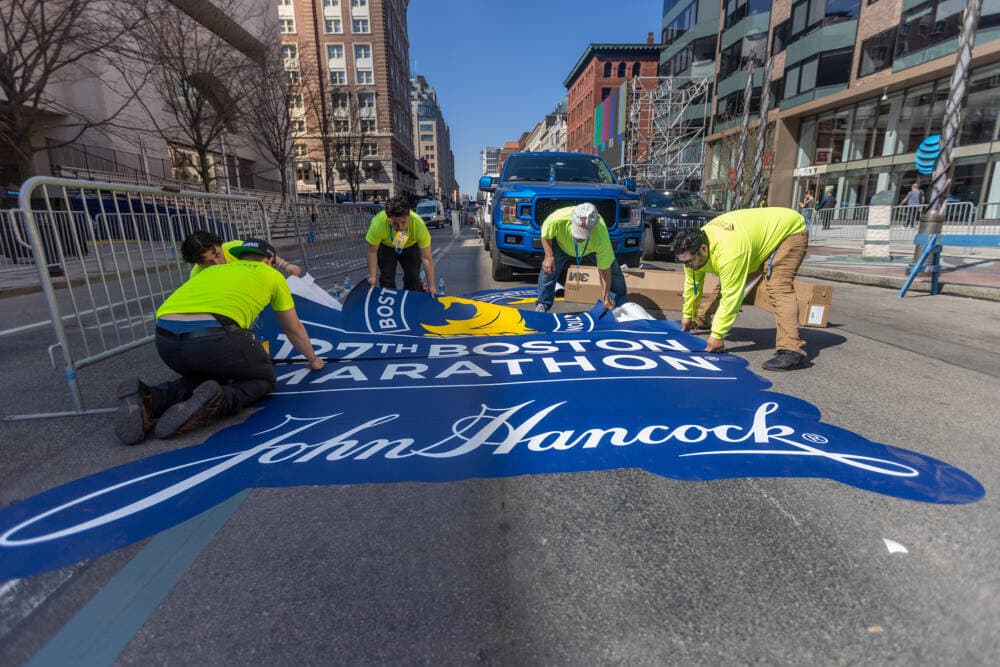 Workers lay down a Boston Marathon decal on Boylston Street with sponsor John Hancock logo, the final time they will sponsor the race. (Jesse Costa/WBUR)