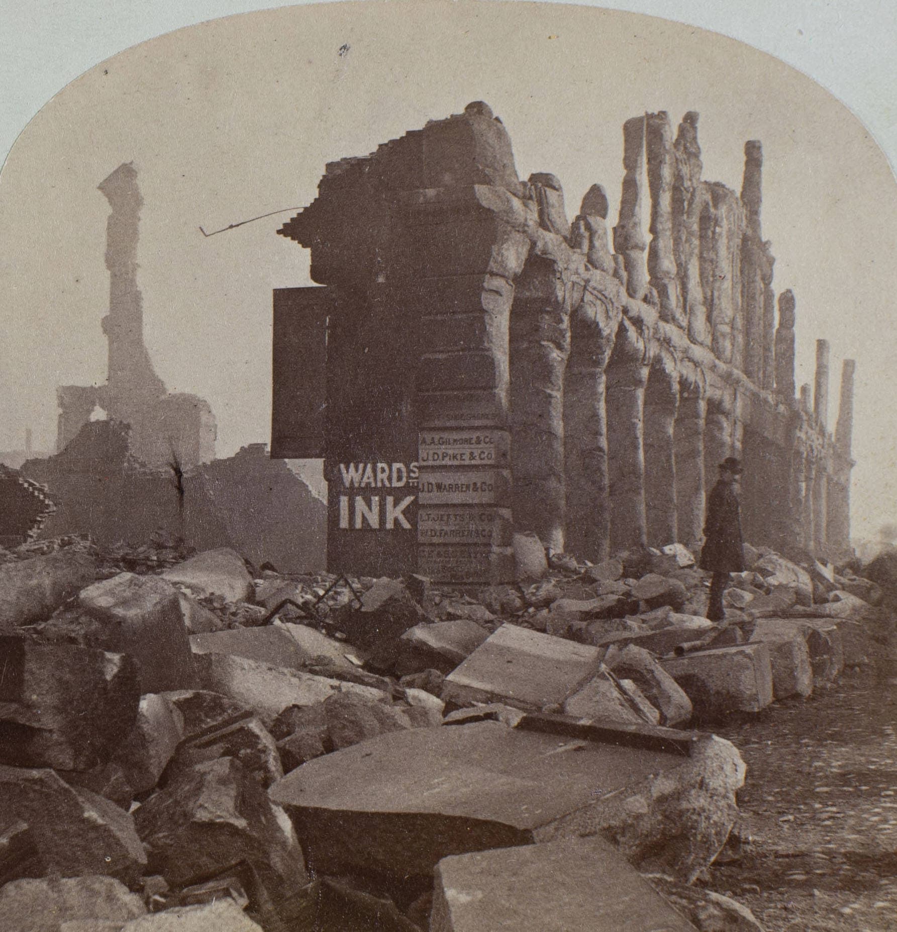 Pearl Street in Boston after the great fire of 1872. Charred partial skeletons of buildings tower over the debris and rubble of their surroundings. A lone man stands on a pile of broken cement blocks next to a facade which is all that remains of a building which, according to a series of signs posted vertically on one of the columns had housed a number of businesses (Courtesy: Boston Athenaeum)