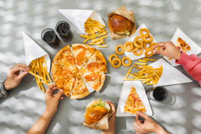Flat-lay of friends group eating burgers, fries, onion rings, pizza, drinking cola at outdoor party over table background, top view. Fast food dinner from delivery service.Fast food and unhealthy eating concept