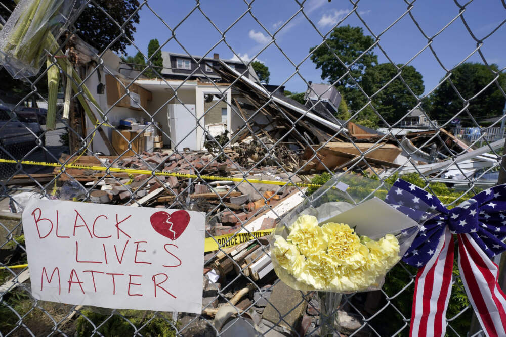 Signs and flowers are attached to a fence outside a building, June 2021, in Winthrop, Mass., where an armed man crashed a hijacked truck on June 26, then fatally shot two Black people before being killed by police. (Steven Senne/AP)