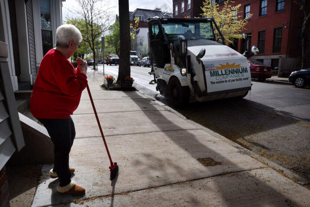 A woman takes a break from sweeping the sidewalk to watch a street sweeper go down Otis Street in Cambridge. A city pilot program eliminates towing during street sweeping. (Craig F. Walker/The Boston Globe via Getty Images)