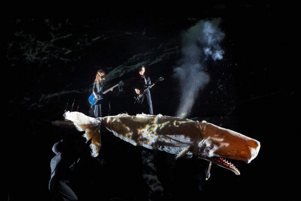 Two musicians play against a black background. A large whale puppet is in the foreground.