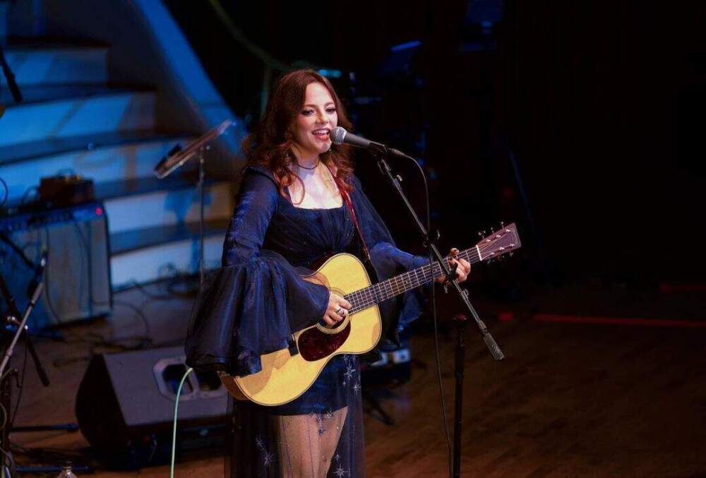 Female singer strumming a guitar on stage in Worcester, Massachusetts. She is wearing a short black dress and a navy blue jacket with bell sleeves.